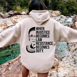 May include: A beige hoodie with a black and white graphic print of Ruth Bader Ginsburg and the text "When injustice becomes law resistance becomes duty."