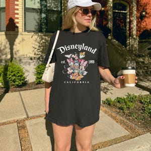 May include: Black t-shirt with the Disneyland logo, the text "est. 1955 CALIFORNIA", and a graphic of Disney characters. The shirt is worn by a person outdoors, with a white purse and sunglasses.