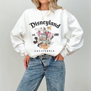 May include: White crew neck sweatshirt with the Disneyland logo and cartoon characters including Mickey Mouse and Donald Duck. The text "est. 1955 CALIFORNIA" is printed below the graphic. The sleeves are long and the fit is relaxed.