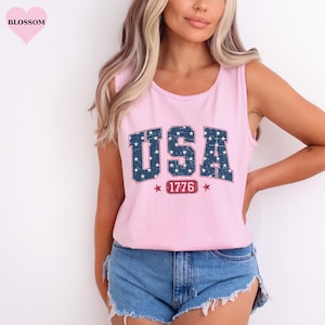 May include: Pink tank top with the word "USA" in a blue, star-filled font. Below the word "USA" is the number "1776" in a red box, with red stars on either side. The tank top is sleeveless.