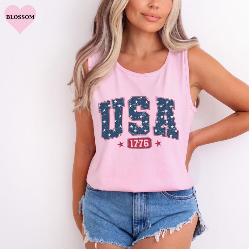 May include: Pink tank top with the word "USA" in a blue, star-filled font. Below the word "USA" is the number "1776" in a red box, with red stars on either side. The tank top is sleeveless.