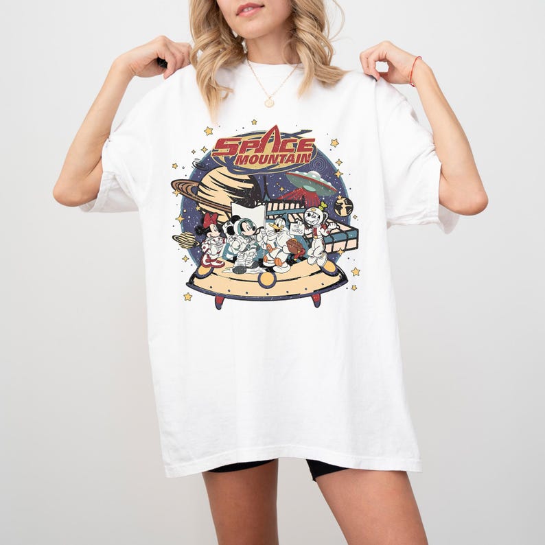 May include: White t-shirt featuring a colorful graphic of Disney characters on a spaceship, with the words "Space Mountain" in a retro font. The design includes planets, stars, and a retro space theme.