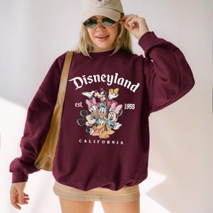 May include: Burgundy sweatshirt featuring the word "Disneyland" in white script above a graphic of Disney characters. The text "est. 1955 CALIFORNIA" is below. The sweatshirt has long sleeves and a crew neck.