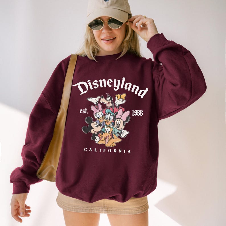 May include: Burgundy sweatshirt featuring the word "Disneyland" in white script above a graphic of Disney characters. The text "est. 1955 CALIFORNIA" is below. The sweatshirt has long sleeves and a crew neck.
