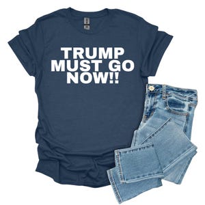Può includere: T-shirt blu navy con la scritta bianca "TRUMP MUST GO NOW!!" e un paio di jeans in denim blu chiaro. La t-shirt è a maniche corte e ha lo scollo tondo. I jeans sono piegati.