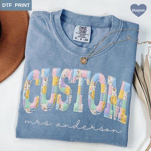 Può includere: Una maglietta blu Comfort Colors con la parola "CUSTOM" in un design patchwork. Sotto la parola "CUSTOM" c'è il nome "mrs. anderson" in corsivo. Una collana d'oro con un ciondolo è drappeggiata sulla maglietta.