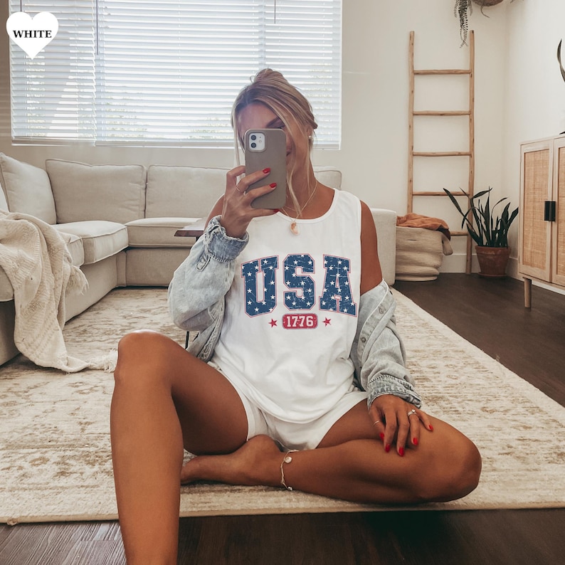 May include: White tank top with the word "USA" in blue letters with white stars, and the number "1776" in red. The tank top is sleeveless and has a round neckline. The tank top is made of a soft, lightweight material.