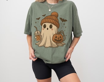 May include: A moss green short-sleeve t-shirt featuring a graphic of a ghost wearing a brown knit beanie, holding an iced coffee, and standing next to a jack-o'-lantern. Small bats and stars are scattered around the central ghost illustration.