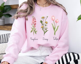 May include: A light pink long-sleeve sweatshirt featuring a graphic print of three flowers with their names written underneath. The design includes a pink foxglove, white daisies, and pink tulips with the text Foxglove, Daisy, and Tulip.