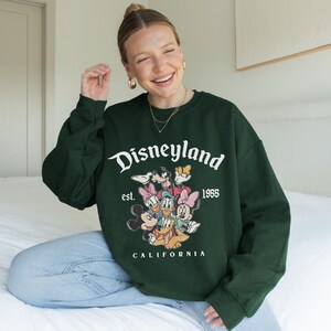 May include: A dark green sweatshirt with the Disneyland logo and the text "est. 1955 CALIFORNIA", featuring Disney characters. The sweatshirt is long-sleeved and has a crew neck.