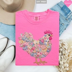 May include: A bright pink t-shirt featuring a colorful floral chicken design. The chicken is made of various fabric patches with a mix of floral patterns. The shirt has a crew neck and the brand name "Comfort Colors" is visible.