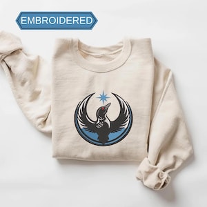 May include: Cream-colored sweatshirt with an embroidered design of a black bird with outstretched wings, a red eye, and a blue star above it. The design is encircled by a blue ring. The word "EMBROIDERED" is in a blue banner at the top.
