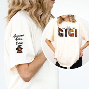May include: An off-white t-shirt featuring the text GIGI on the front with Halloween-themed patterns including a spiderweb, bats, and pumpkins. The sleeve displays the names Alexander, Olivia, and Sarah above a graphic of a pumpkin wearing a witch hat.