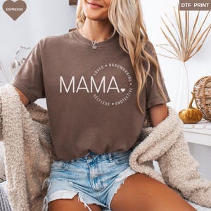 May include: Espresso-colored t-shirt with the word "MAMA" in white, encircled by "LOVED, HARDWORKING, SELFLESS, PROTECTIVE." The shirt is paired with denim shorts and a cream-colored cardigan. The image also includes a decorative pumpkin and a woven basket.