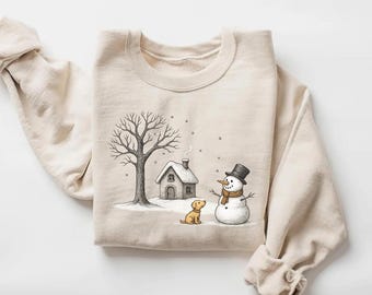 May include: Cream-colored sweatshirt featuring a winter scene illustration. The design includes a snowman wearing a top hat and scarf, a small house, a bare tree, and a dog. Snowflakes are falling in the background.