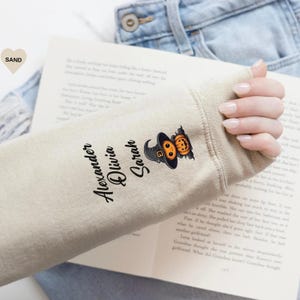 May include: A sand-colored long-sleeve shirt sleeve featuring the printed names Alexander, Olivia, and Sarah in a black script font, accompanied by a graphic of a small orange pumpkin wearing a black witch hat with a gold buckle.
