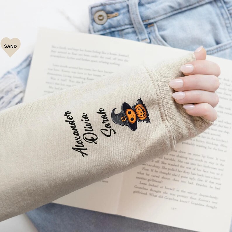 May include: A sand-colored long-sleeve shirt sleeve featuring the printed names Alexander, Olivia, and Sarah in a black script font, accompanied by a graphic of a small orange pumpkin wearing a black witch hat with a gold buckle.