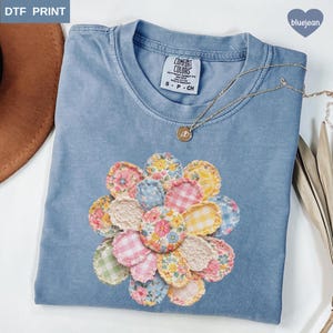 May include: A light blue Comfort Colors t-shirt with a floral design. The design is made of various fabric patterns in pink, yellow, blue, and green. A gold necklace with a pendant is draped on the shirt.