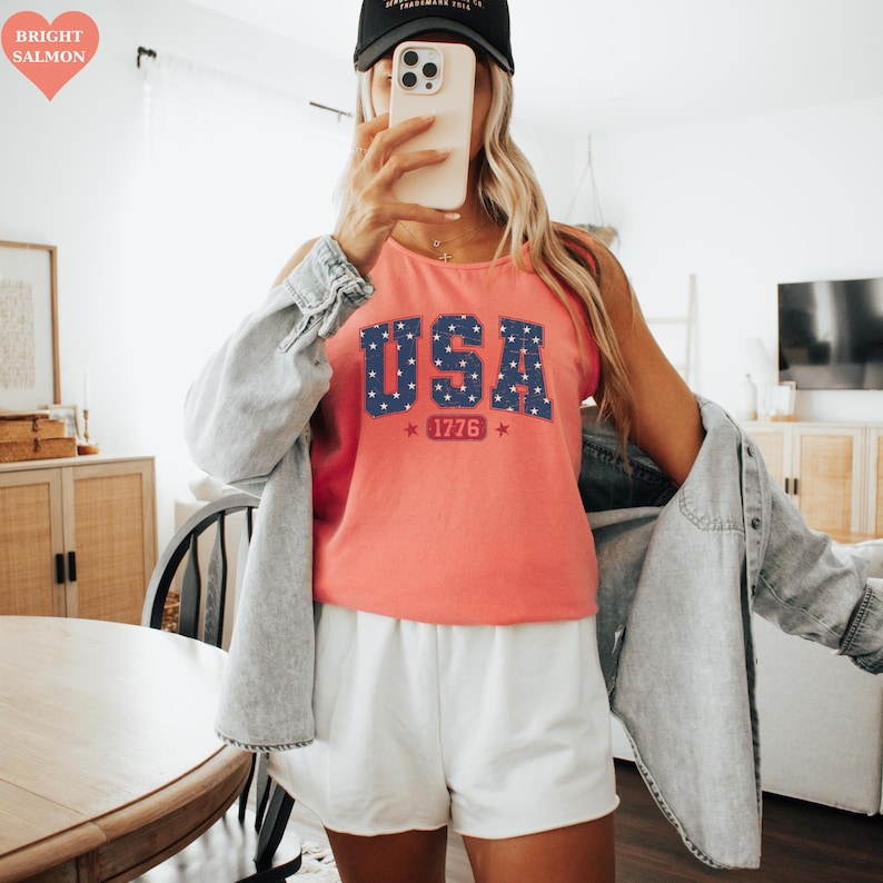 May include: A coral-colored tank top with the word "USA" in navy blue, star-filled letters. Below the text is "1776" in red. The tank top is paired with white shorts and a light blue denim jacket.