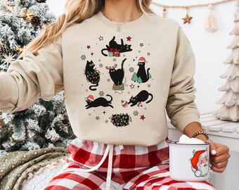 May include: A beige sweatshirt with a festive design of black cats wearing Santa hats, Christmas lights, and gifts. The cats are surrounded by stars and decorative elements, creating a holiday theme. The sweatshirt is paired with red and white plaid pajama bottoms.