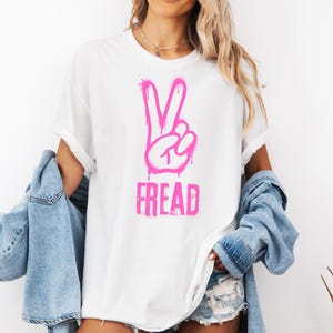 Puede incluir: Camiseta blanca con un signo de la paz rosa y la palabra "FREAD" en estilo grafiti. La camiseta tiene mangas enrolladas y un corte holgado.