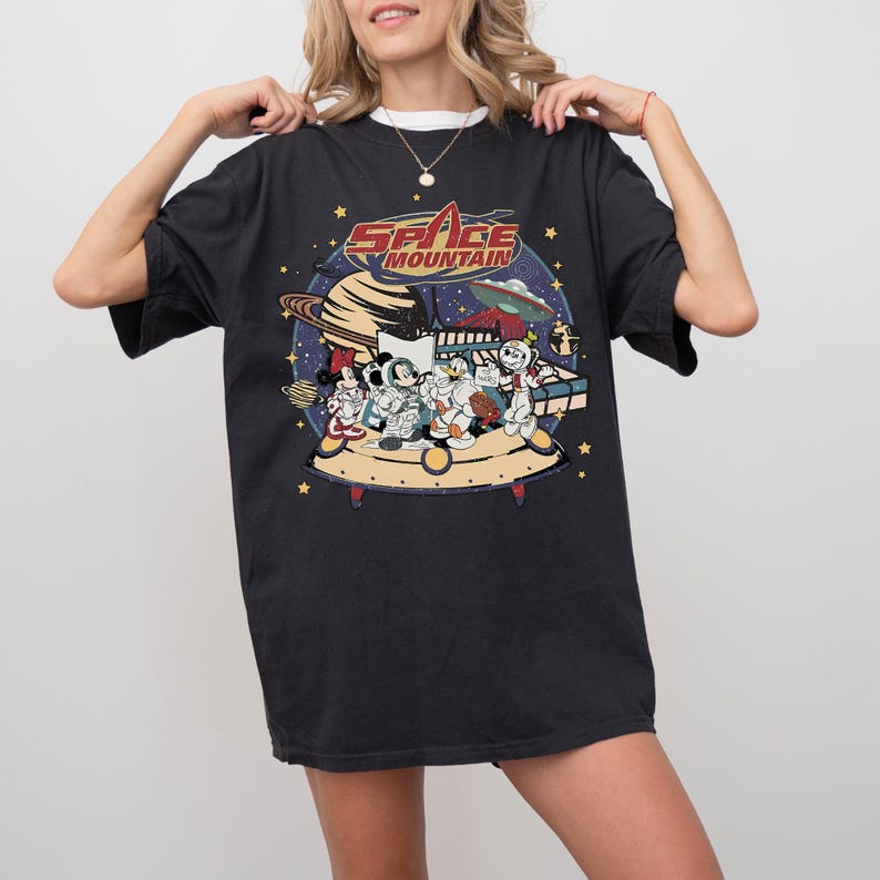 May include: Black t-shirt featuring a colorful graphic of cartoon characters on a spaceship, with the words "Space Mountain" in red and white. The design includes planets, stars, and a UFO, creating a space-themed illustration.