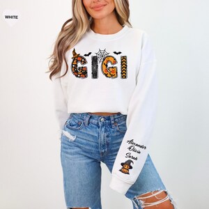 May include: A white long-sleeve sweatshirt featuring the text GIGI in a Halloween-themed pattern with pumpkins, bats, and a spiderweb. The left sleeve displays the names Alexander, Olivia, and Sarah above a small illustration of a pumpkin witch.