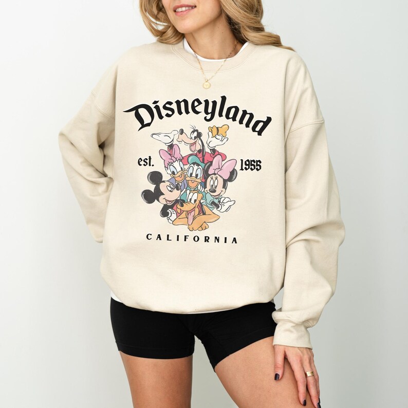 May include: Beige sweatshirt with a Disneyland graphic featuring cartoon characters. The text reads "Disneyland est. 1955 California" in black. The sweatshirt has long sleeves and a crew neck. The design is a classic.
