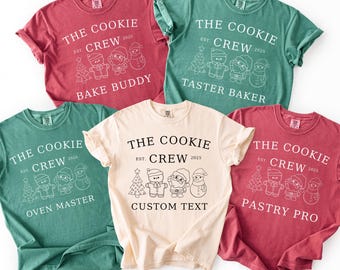 May include: A collection of five short-sleeved t-shirts in various colors: red, green, and cream. Each shirt features the text "THE COOKIE CREW EST. 2025" with different titles below, such as "BAKE BUDDY" and "CUSTOM TEXT".