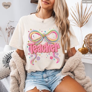 May include: Ivory-colored t-shirt featuring a colorful bow design with the word "Teacher" in pink, glittery letters. The bow has rainbow-colored stripes and gold glitter accents. The shirt has a relaxed fit and short sleeves.