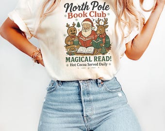 Puede incluir: Camiseta color crema con un gráfico de Papá Noel, un hombre de jengibre y un reno leyendo libros. El texto dice "North Pole Book Club" y "Magical Read! Hot Cocoa Served Daily".