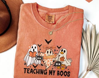 May include: A terracotta-colored t-shirt featuring a graphic of three ghosts, one holding a book, one with a bow holding a pencil, and one holding a pumpkin, surrounded by spiders and bats, with the text "TEACHING MY BOOS" printed below.