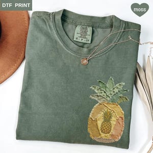 May include: A moss green t-shirt with a pineapple applique. The pineapple design features a detailed illustration in the center, surrounded by patches of yellow, orange, and green fabric. A gold necklace with a pendant rests on the shirt.