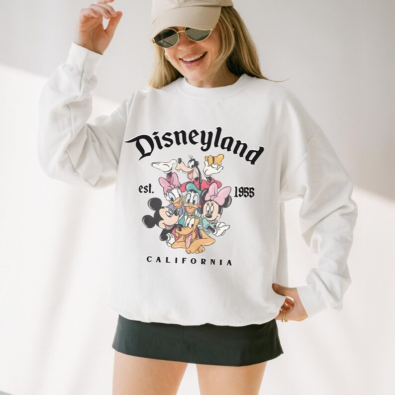 May include: White sweatshirt with the Disneyland logo and cartoon characters Mickey Mouse, Minnie Mouse, Donald Duck, Goofy, and Daisy Duck. The text "est. 1955 CALIFORNIA" is also printed on the sweatshirt. A classic design.