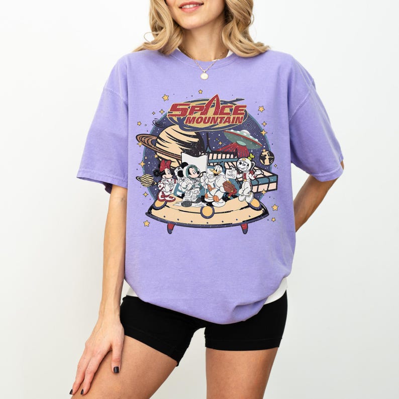 May include: A light purple t-shirt featuring a graphic of Disney characters on a spaceship with the words "Space Mountain". The design includes planets, stars, and an alien spaceship. The shirt has short sleeves and a crew neck.