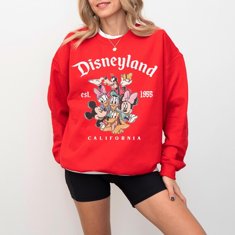 May include: A red Disneyland sweatshirt with a graphic of Disney characters including Mickey and Minnie Mouse. The sweatshirt has the text "Disneyland" in white, with "est. 1955" and "CALIFORNIA" below the graphic.