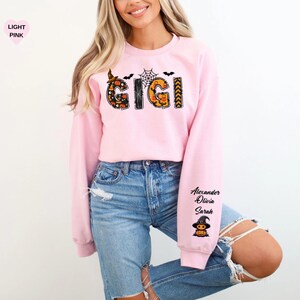 May include: A light pink long-sleeve sweatshirt featuring the word GIGI in a Halloween-themed pattern with pumpkins, spiderwebs, and bats. The left sleeve displays the names Alexander, Olivia, and Sarah above a small graphic of a witch pumpkin.