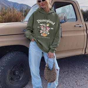 May include: An olive green Disneyland hoodie featuring Mickey Mouse, Minnie Mouse, Donald Duck, Daisy Duck, and Goofy. The hoodie has the text "Disneyland" and "California". The person is wearing light blue jeans and carrying a leopard print handbag.