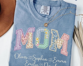 May include: A light blue t-shirt with the word "MOM" appliqued in colorful floral and patterned fabrics. Below the word "MOM" are the names "Oliver", "Sophia", "Emma", "Emily", and "David". A gold necklace is draped across the shirt.