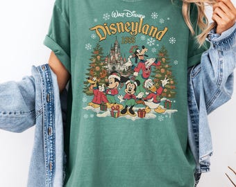 May include: Green t-shirt featuring a Disneyland 1955 graphic with Disney characters in festive attire. The design includes Mickey Mouse, Minnie Mouse, Donald Duck, Goofy, and Pluto, with a castle and Christmas trees. The shirt has a crew neck and short sleeves.