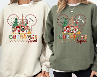 May include: Two sweatshirts, one beige and one olive green, featuring a Christmas-themed graphic. The design includes Mickey Mouse ears, a castle, Christmas trees, and the words "Christmas Squad". The beige sweatshirt has the name "Sarah" and the green one has "Custom".