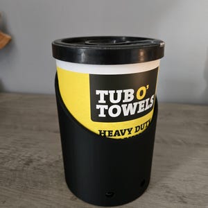 May include: A black plastic container with a yellow label that says "TUBO' TOWELS HEAVY DUTY" on it. The container is sitting on a wooden surface.