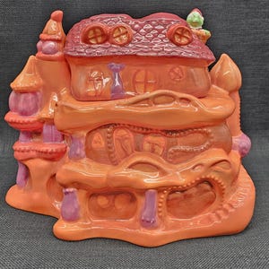 May include: A whimsical, ceramic house sculpture in shades of orange, pink, and red. The multi-tiered structure features rounded windows, a textured roof, and decorative accents. The overall design is reminiscent of a fairytale cottage.