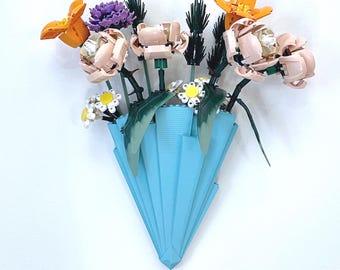 May include: A light blue origami vase displays a vibrant bouquet of flowers. The arrangement features orange, purple, and cream-coloured blooms, with green leaves and stems. The vase is mounted on a white wall, presenting a unique floral display.