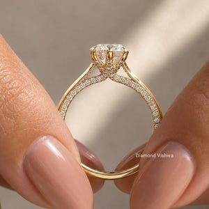 May include: A gold ring with a large, clear gemstone at the center. The band is adorned with small, sparkling stones. The ring is held between two hands with manicured nails. The ring is a piece of jewelry.