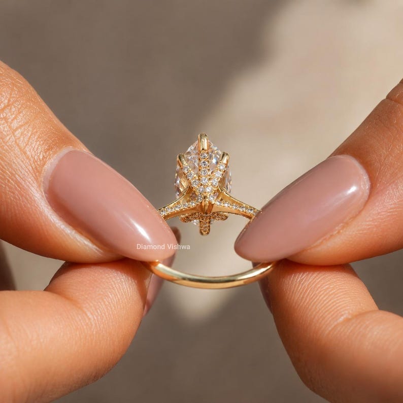 May include: A gold ring with a pear-shaped diamond center stone. The ring features a delicate band and intricate detailing around the diamond. The ring is held between two hands. The text "Diamond Vishwa" is visible.