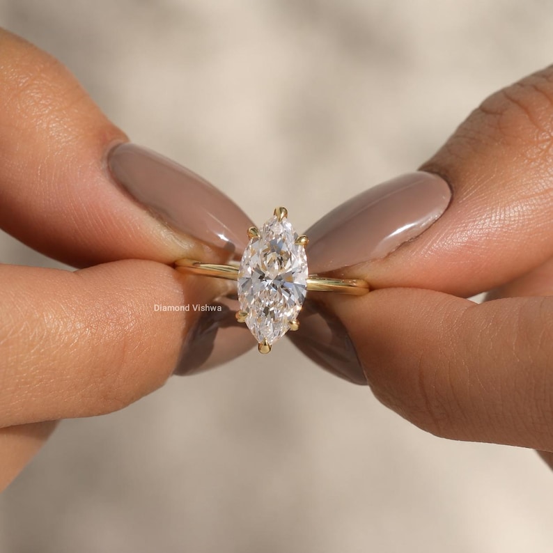 May include: A marquise-cut diamond ring with a slender gold band. The diamond is set with prongs and sparkles brightly. The ring is held between two hands with manicured nails. The text "Diamond Vishwa" is visible.