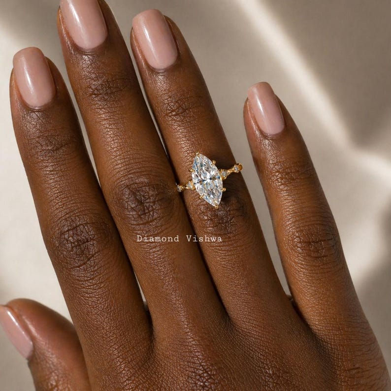 May include: A gold ring with a large, marquise-cut diamond as the centerpiece. The ring has smaller diamonds on the band and is set against a dark skin tone. The text "Diamond Vishwa" is visible.