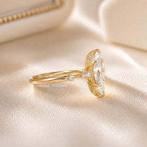 May include: A gold ring with a large, oval-shaped clear stone. The ring has a delicate band with small accent stones and intricate detailing around the main stone. The text "Diamond Vishwa" is visible on the band.