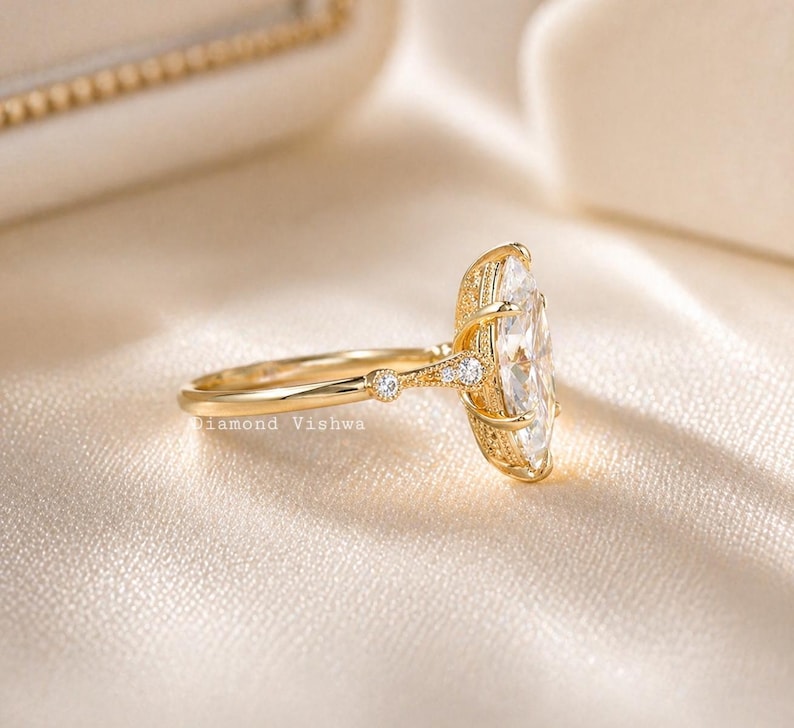 May include: A gold ring with a large, oval-shaped clear stone. The ring has a delicate band with small accent stones and intricate detailing around the main stone. The text "Diamond Vishwa" is visible on the band.